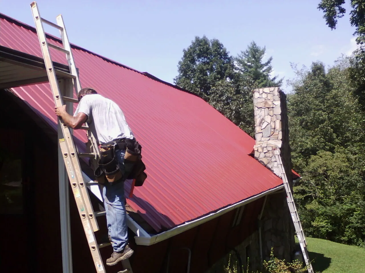 Roofer on a ladder beside a red metal roof and stone chimney — Soffit & Fascia Repair by Roofing Brothers in Cummaquid